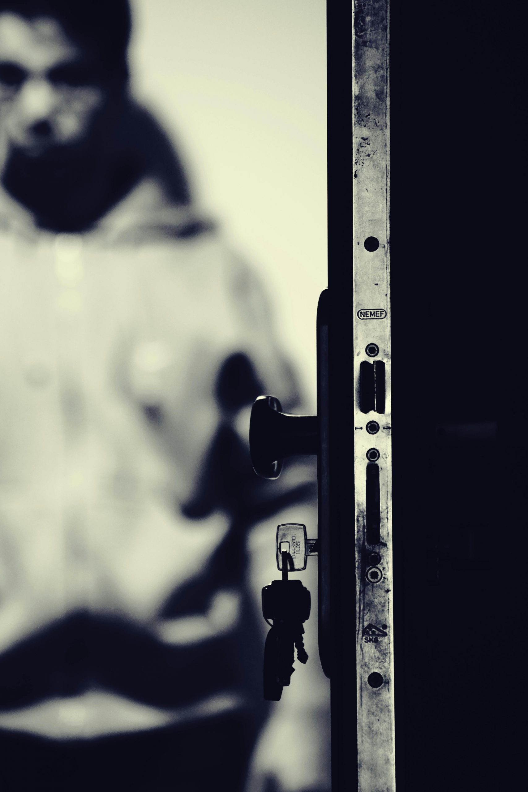 A blurred figure stands behind an open door, focus on lock and keys.